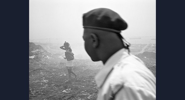 Conflict between Luanda’s population and its governing elites forms an undercurrent to this photograph of a young woman carrying a baby across litter-strewn ground, observed by a man wearing a military beret. 